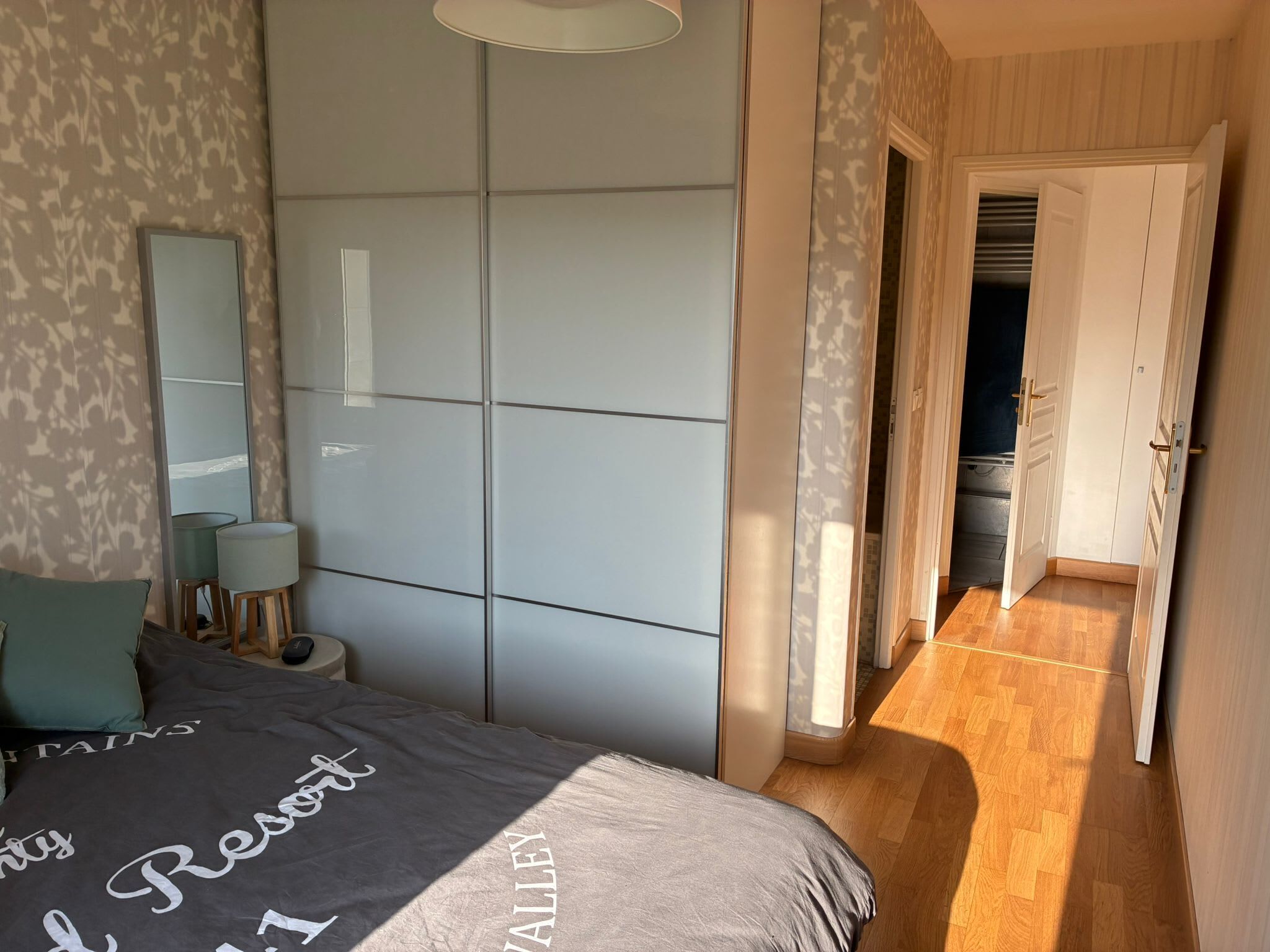 A bedroom with a bed, bedside lamps, a wardrobe, and a view of a hallway leading to a door.