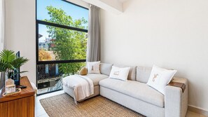 Un salon moderne avec un canapé, une table basse et une grande fenêtre donnant sur des arbres et des immeubles.
