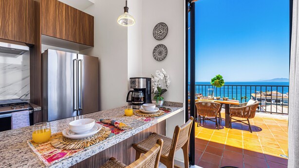 A kitchen with a refrigerator, coffee maker, and dining area with chairs and a table. - Huge Private Terrace - Panoramic Bay Views - Walk to the Malecón (Puerto Vallarta)
