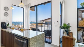 Une cuisine moderne dotée d’un grand îlot central, d’un coin repas avec des chaises et offrant une vue sur la mer grâce à de grandes fenêtres.