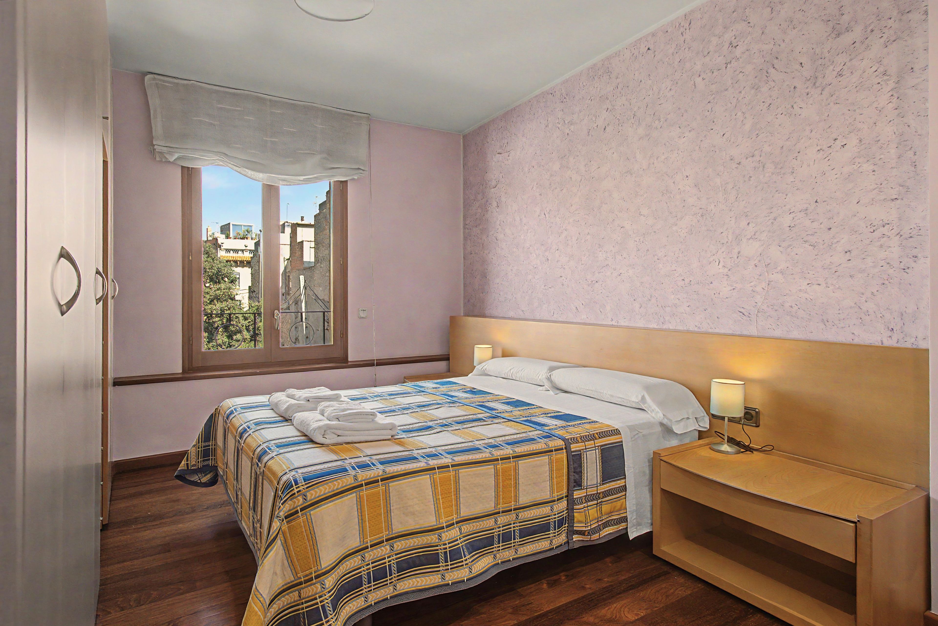 A bedroom with a bed, bedside table, lamp, window, and view of buildings.