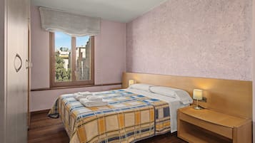 A bedroom with a bed, bedside table, lamp, window, and view of buildings.