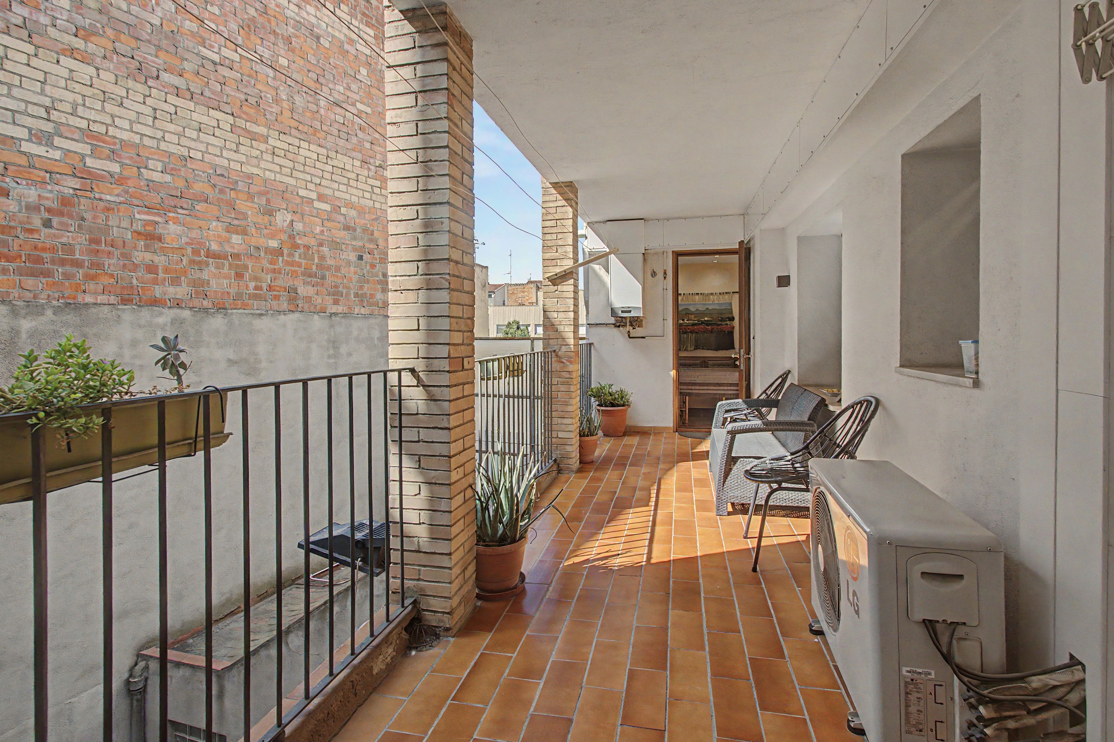 A narrow outdoor corridor with a tiled floor, a brick wall on one side, and a white wall on the other. There is a metal railing on the left, a potted plant, and a table with chairs on the right.