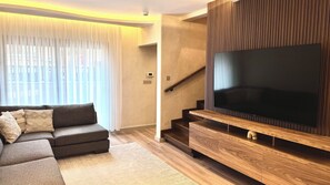 A modern living room with a sectional sofa, a flat-screen TV mounted on a wooden panel, and a staircase leading to an upper level.