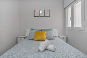 A neatly made bed with patterned bedding, a single yellow pillow, and a rolled white towel. There are two bedside tables with drawers, and a window with blinds on the right. - Triviño Granada Centro Apartment with Wi-Fi, Air Conditioning and Optional Parking (Granada)