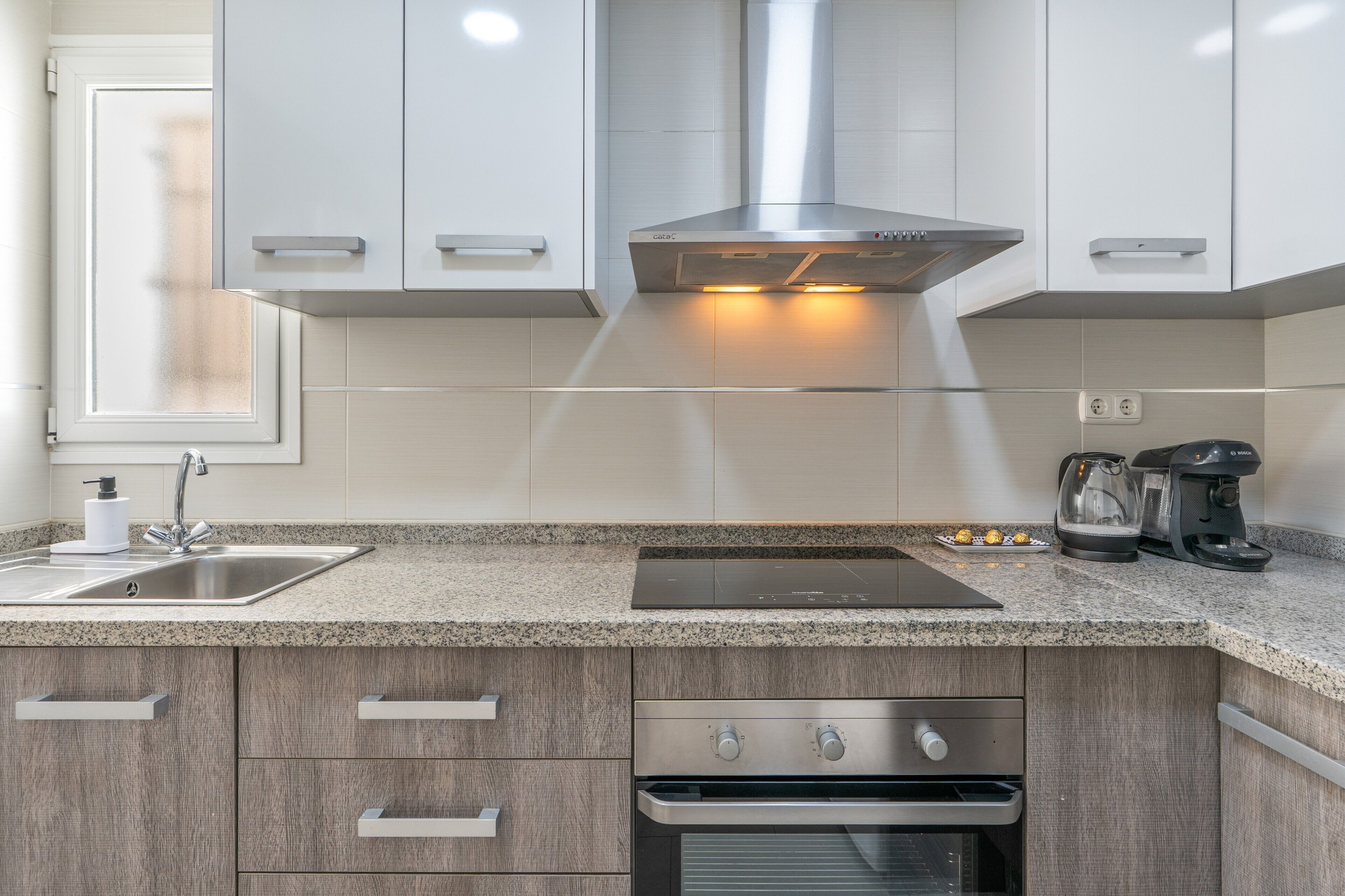 A modern kitchen with a stainless steel range hood, a built-in oven, a coffee maker, and a sink.