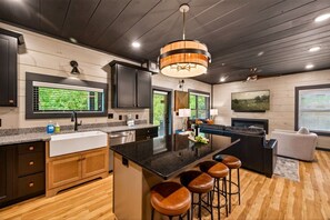 A modern kitchen with a central island, bar stools, and a dining area with a sofa and a coffee table. - Next Play Clubhouse | Waterpark Resort & Game Room (Sevierville)
