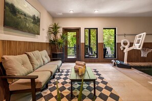 Ein Wohnzimmer mit einer Couch, einem Couchtisch mit Getränken, einem Basketballkorb und Blick auf Grün hinter großen Fenstern.