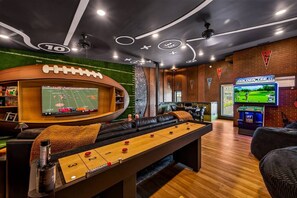 Une table de billard, une table de baby-foot et un écran de télévision diffusant un match de football se trouvent dans un espace de détente aux murs en briques et au sol recouvert d’un tapis vert.