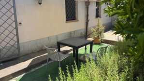 Et udendørs opholdsområde med et bord, stole, en potteplante og et vindue med et metalgitter.