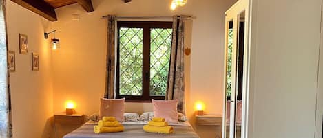 A bedroom with a wooden ceiling, a bed with pillows and a blanket, a nightstand with a lamp, a window with curtains, and a wardrobe.