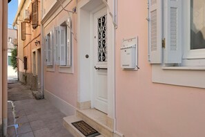 A narrow street with a pink building featuring a white door with a decorative metal grille, a small step leading to the entrance, and a utility box on the right side. - Casabella Apartment in Corfu Town with Air Conditioning (Corfu)