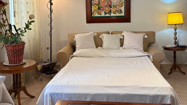 Quarto com cama, pintura, luminária de chão, mesa com pratos decorativos e planta em vaso.