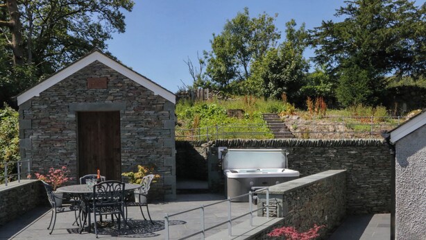 A stone building with a door, a hot tub, and a patio with chairs and a table. - Cragg Cottage (Coniston)