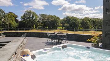 A hot tub with a stone wall and a table with chairs, surrounded by a stone deck and a view of a grassy field.
