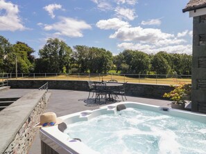 A hot tub with a stone wall and a table with chairs, surrounded by a stone deck and a view of a grassy field. - Cragg Cottage (Coniston)