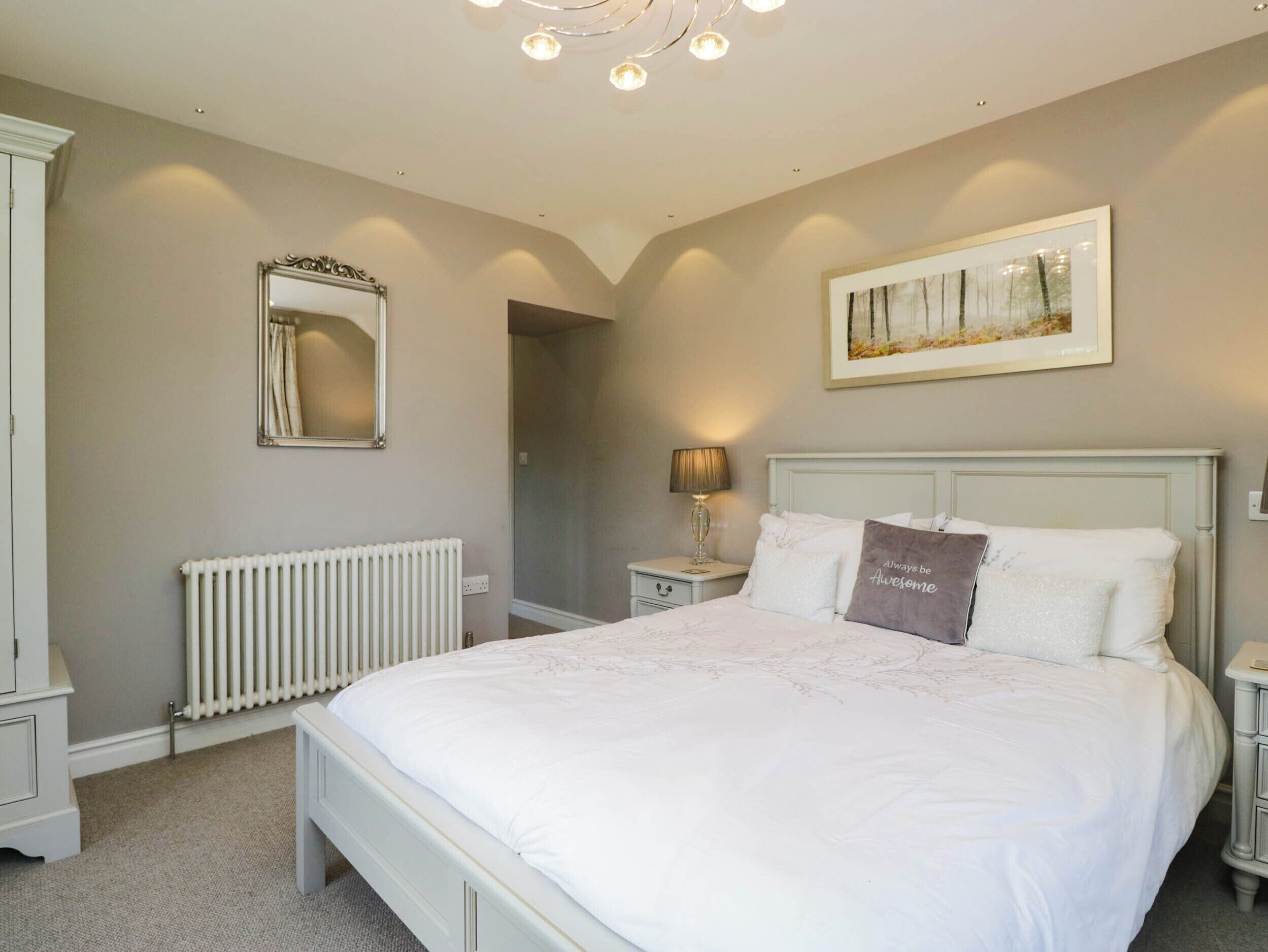 A bedroom with a large bed, a nightstand, a lamp, a radiator, a mirror, and a framed picture on the wall.