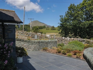 A stone wall with a paved area in front, a stone building, and a view of a hill and trees. - Cragg Cottage (Coniston)