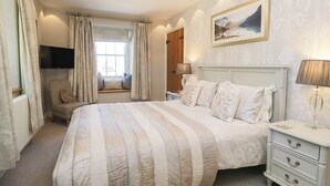 A bedroom with a large bed, a bedside table with a lamp, a television mounted on the wall, and a window with curtains. - Cragg Cottage (Coniston)