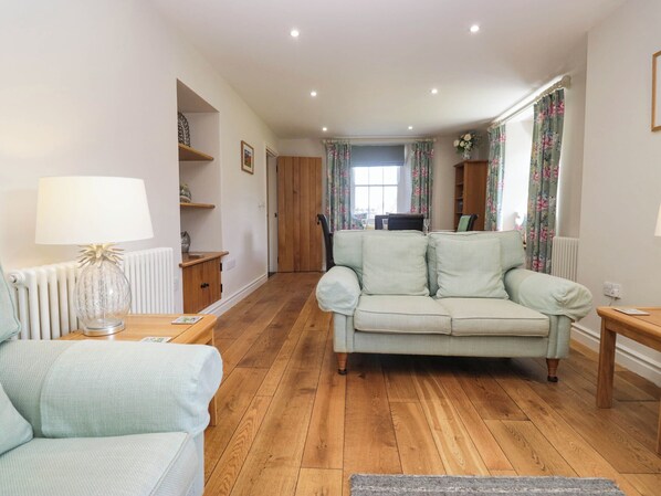A living room with a sofa, a table, and a lamp. There is a radiator and a window with curtains. - Cragg Cottage (Coniston)