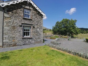 A stone building with two windows, a paved driveway, and a grassy area. - Cragg Cottage (Coniston)
