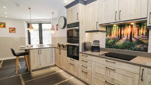 A modern kitchen with wooden cabinets, a built-in oven, a sink, and a decorative forest mural. - Cragg Cottage (Coniston)