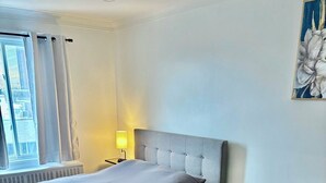 Ett sovrum med en säng, ett sängbord, en lampa, ett element, en byrå och ett fönster med gardiner.