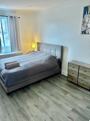 Ein Schlafzimmer mit Bett, Nachttisch, Lampe, Heizkörper, Kommode und einem Fenster mit Vorhängen.