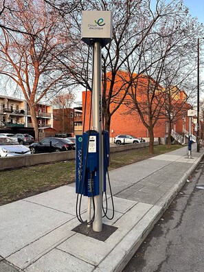 En laddstation för elfordon med en skylt, placerad på en trottoar med träd och byggnader i bakgrunden.
