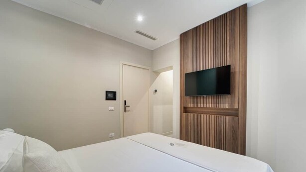 A hotel room with a bed, a television mounted on a wooden panel wall, and a door with a light switch. - Deluxe Double Room (Rome)