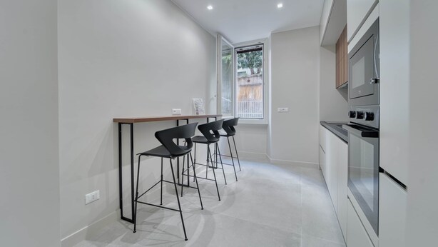 A modern kitchen with a bar area, two bar stools, a built-in oven, and a window with a view of the outside.