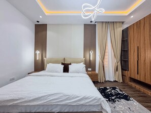 A modern bedroom with a bed, bedside tables, a nightstand, a wall-mounted light fixture, a window with curtains, and a wooden wardrobe. - Exclusive 1-Bedroom in Victoria Island with Pool, Fast Wi-Fi & 24/7 Power (Lagos)