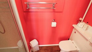 Une salle de bain avec un mur rouge, des toilettes blanches, une petite poubelle et un espace douche.