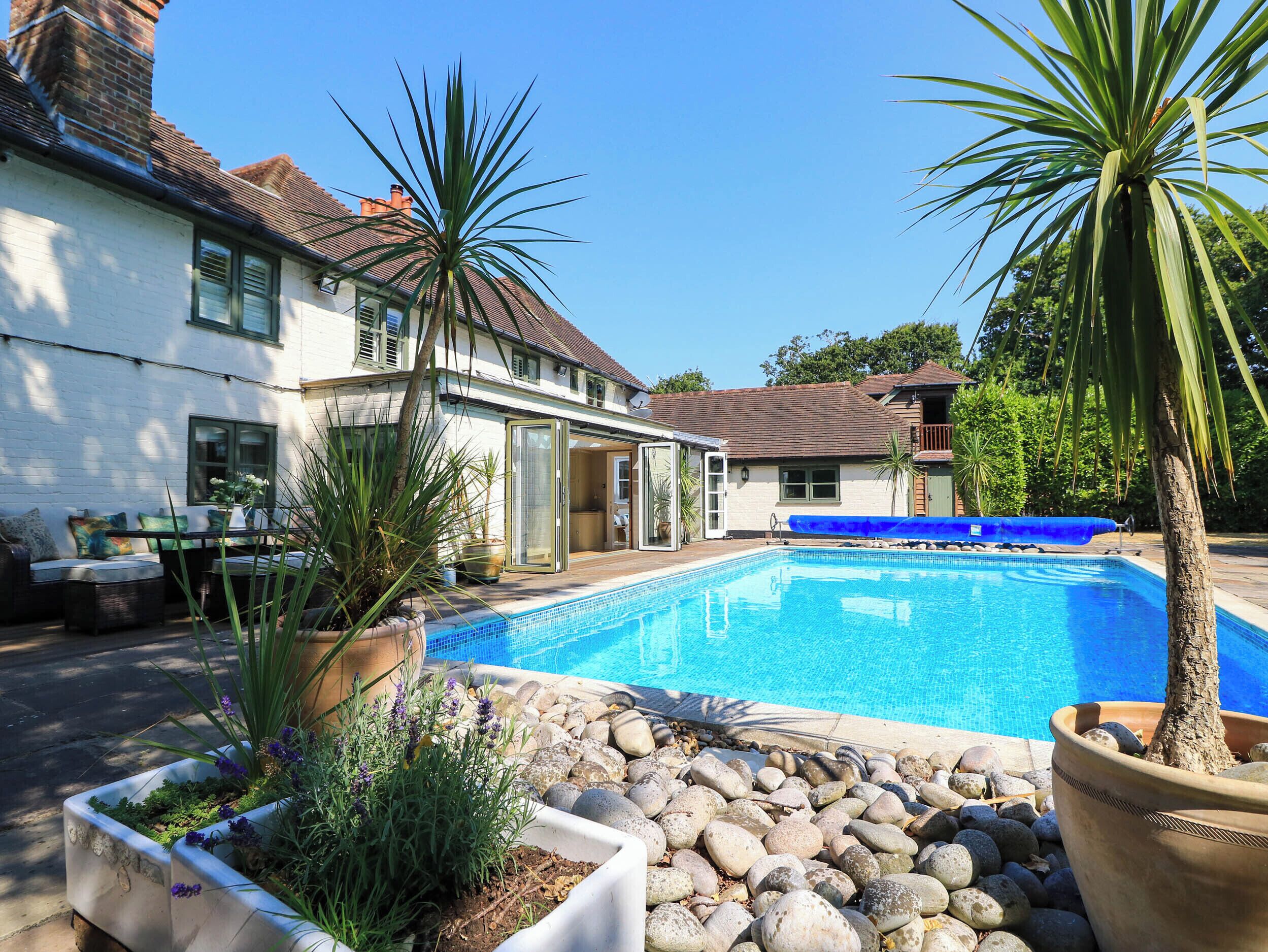 A residential swimming pool with a patio area, surrounded by potted plants and a white house with a brown roof.