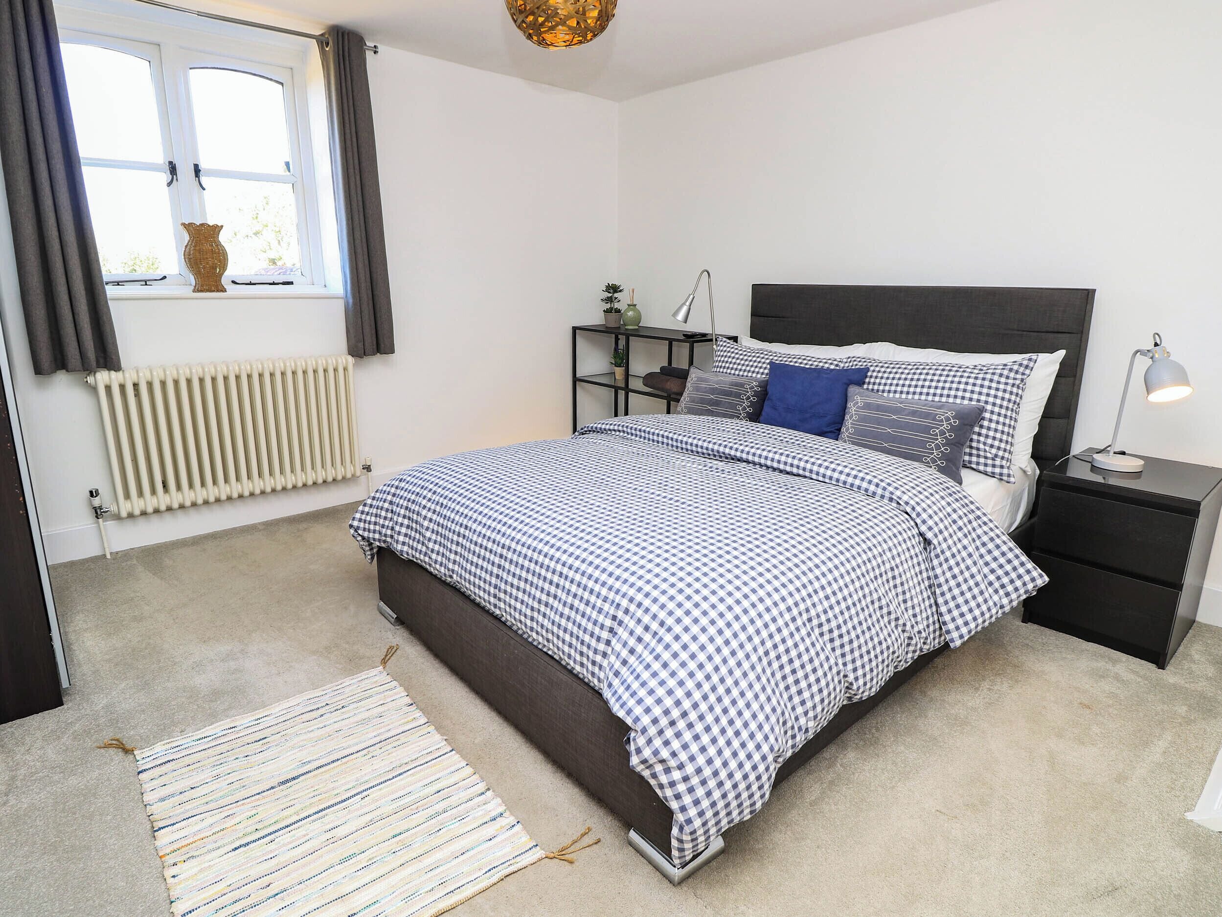 A bedroom with a bed, bedside table, lamp, radiator, window, and curtains.