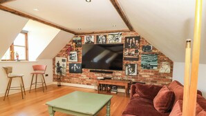 Una habitación estilo loft con techo inclinado, vigas de madera, pared de ladrillo, televisor de pantalla plana, una mesa verde, un sofá rojo y dos taburetes altos.
