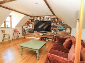 A loft-style room with a sloped ceiling, wooden beams, a brick wall, a flat-screen TV, a green table, a red sofa, and two bar stools.