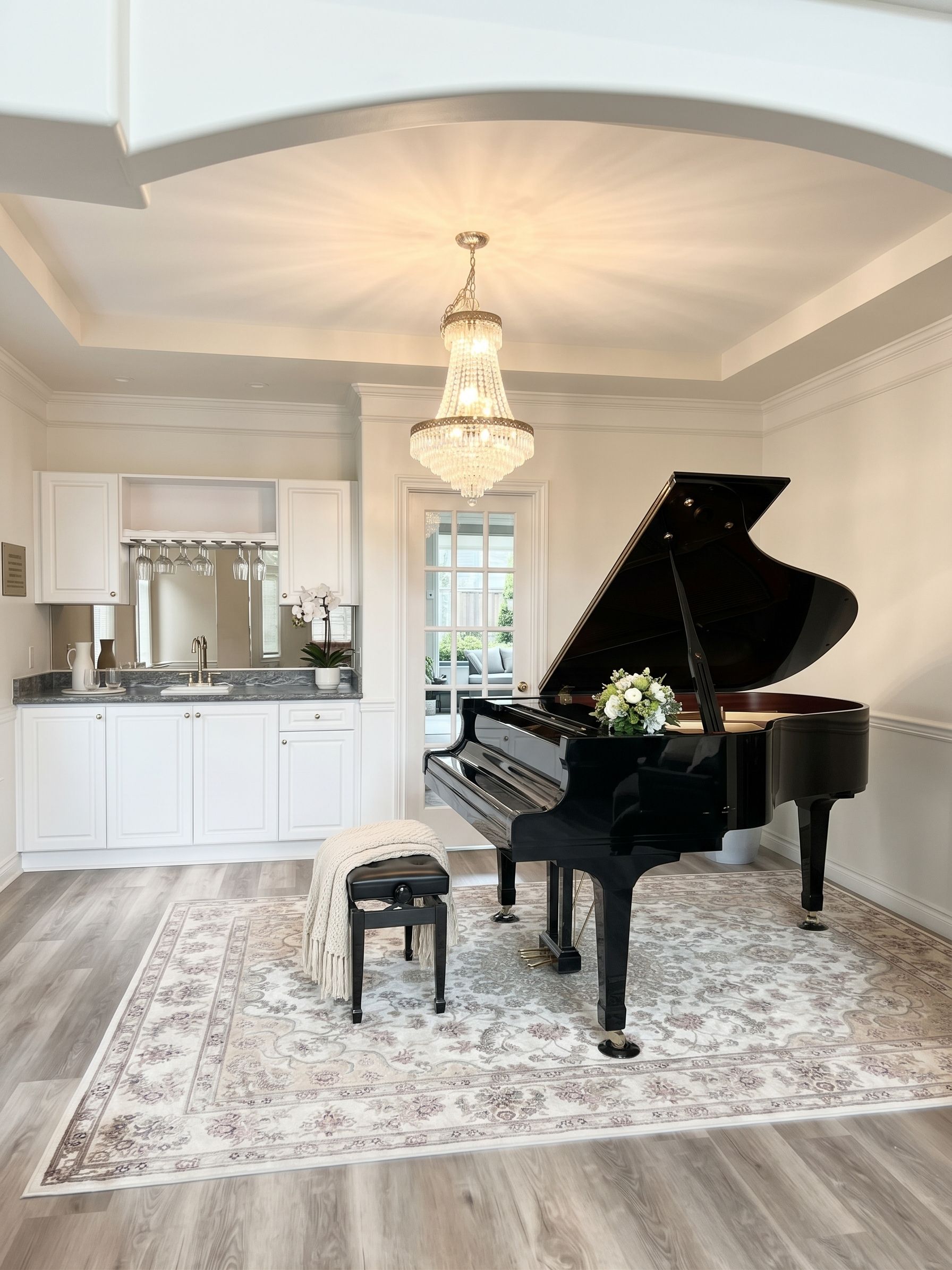 A grand piano in a spacious room with a marble floor, a large chandelier, and a kitchen area with white cabinetry.