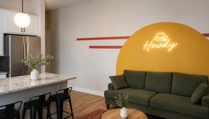 Une cuisine moderne dotée d’un réfrigérateur encastré, un coin repas avec un plan de travail en marbre et un salon avec un canapé vert et un néon.