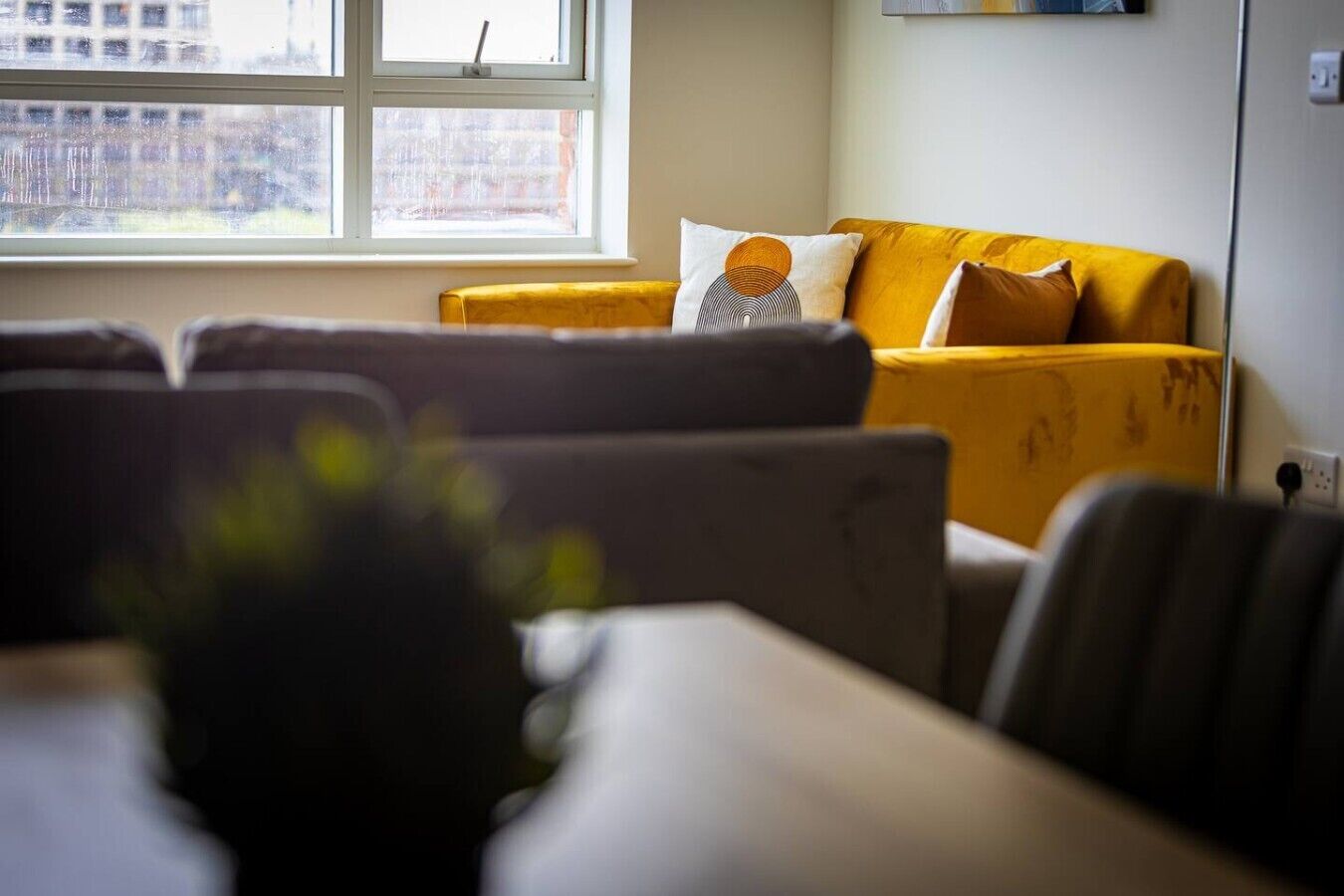 A living room with a sofa and armchairs, a window with a view of buildings, and a wall-mounted shelf.