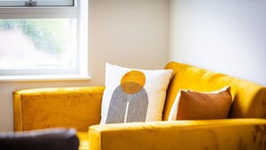 Ein gelbes Sofa mit gemusterten Kissen, ein Fenster mit Blick auf Grünflächen und ein Teil eines dunkel gefärbten Stuhls.