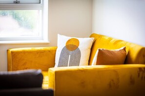 A yellow sofa with patterned cushions, a window with a view of greenery, and a partial view of a dark-colored chair. - Birmingham City Retreat No 146 (Birmingham)