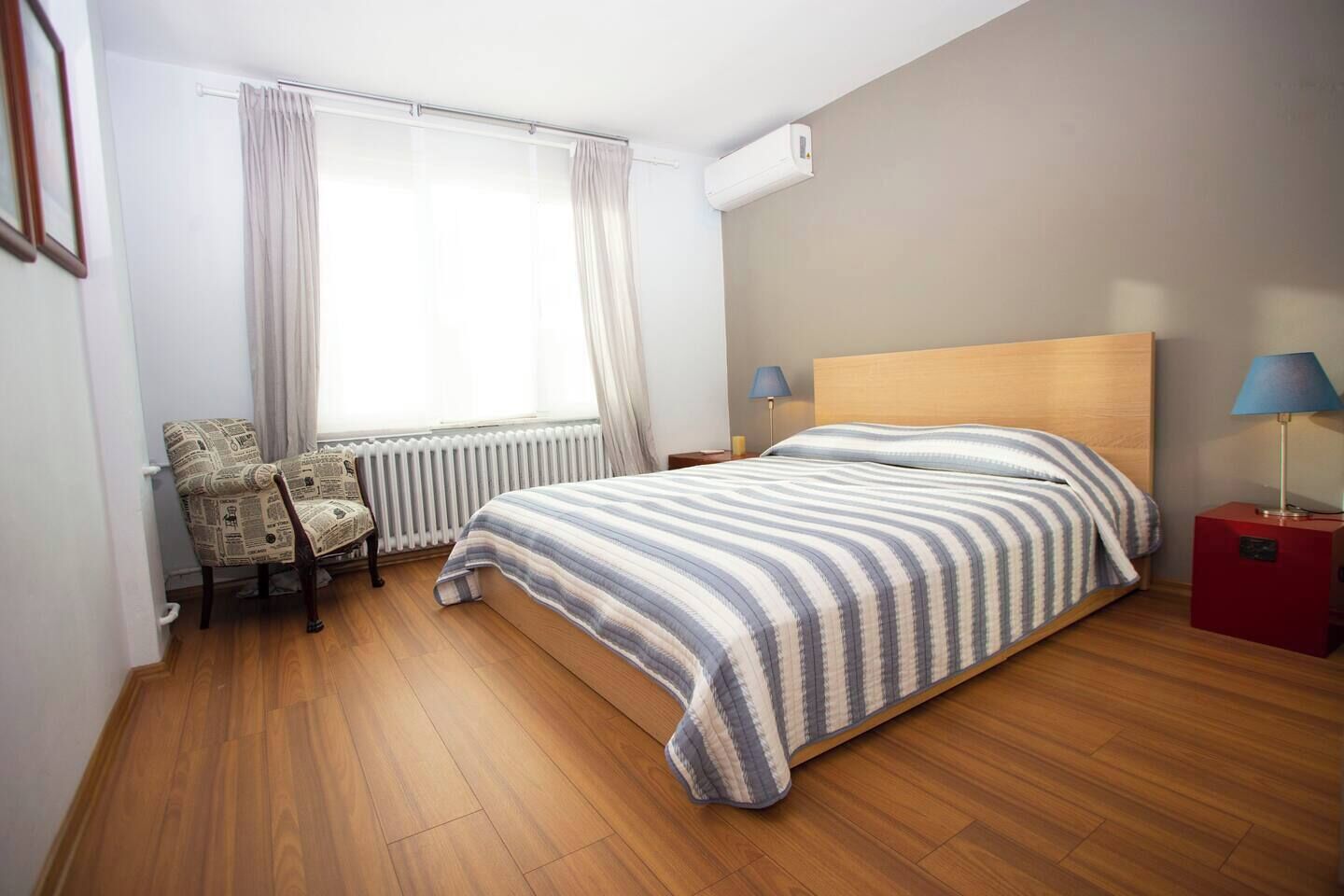 Een slaapkamer met een houten vloer, een bed met gestreepte lakens, een stoel met een krantenprint, een radiator en twee bedlampjes.
