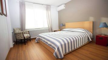 Een slaapkamer met een houten vloer, een bed met gestreepte lakens, een stoel met een krantenprint, een radiator en twee bedlampjes.
