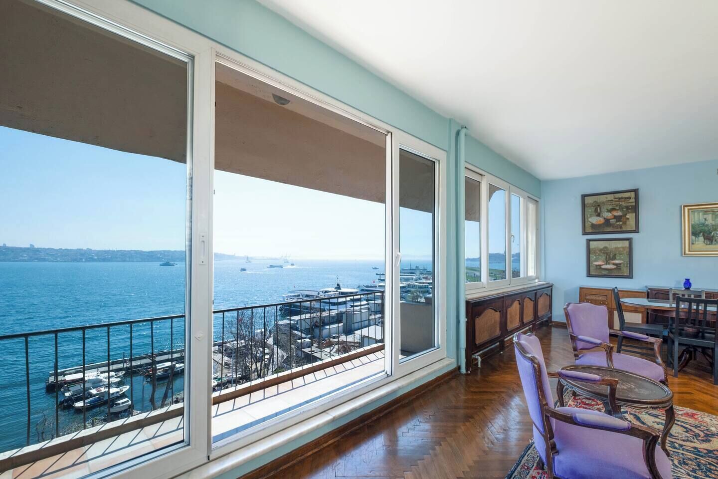 Ein Balkon mit Blick auf das Meer und Boote, Holzboden sowie eine Essecke mit Stühlen und einem Tisch.