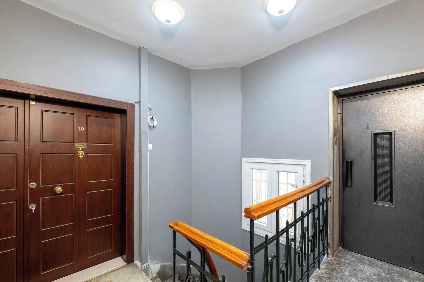 Un couloir avec une porte en bois portant le numéro « 10 », un mur gris, une fenêtre et une porte noire à la surface texturée.