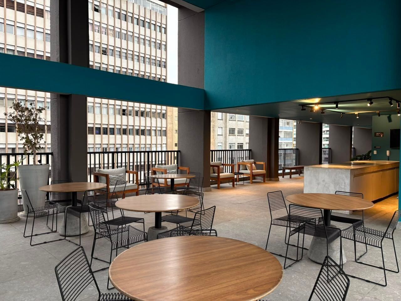 Uma área de estar interna moderna, com mesas e cadeiras redondas, um bar e grandes janelas que oferecem vista para a cidade.