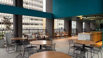 Uma área de estar interna moderna, com mesas e cadeiras redondas, um bar e grandes janelas que oferecem vista para a cidade.