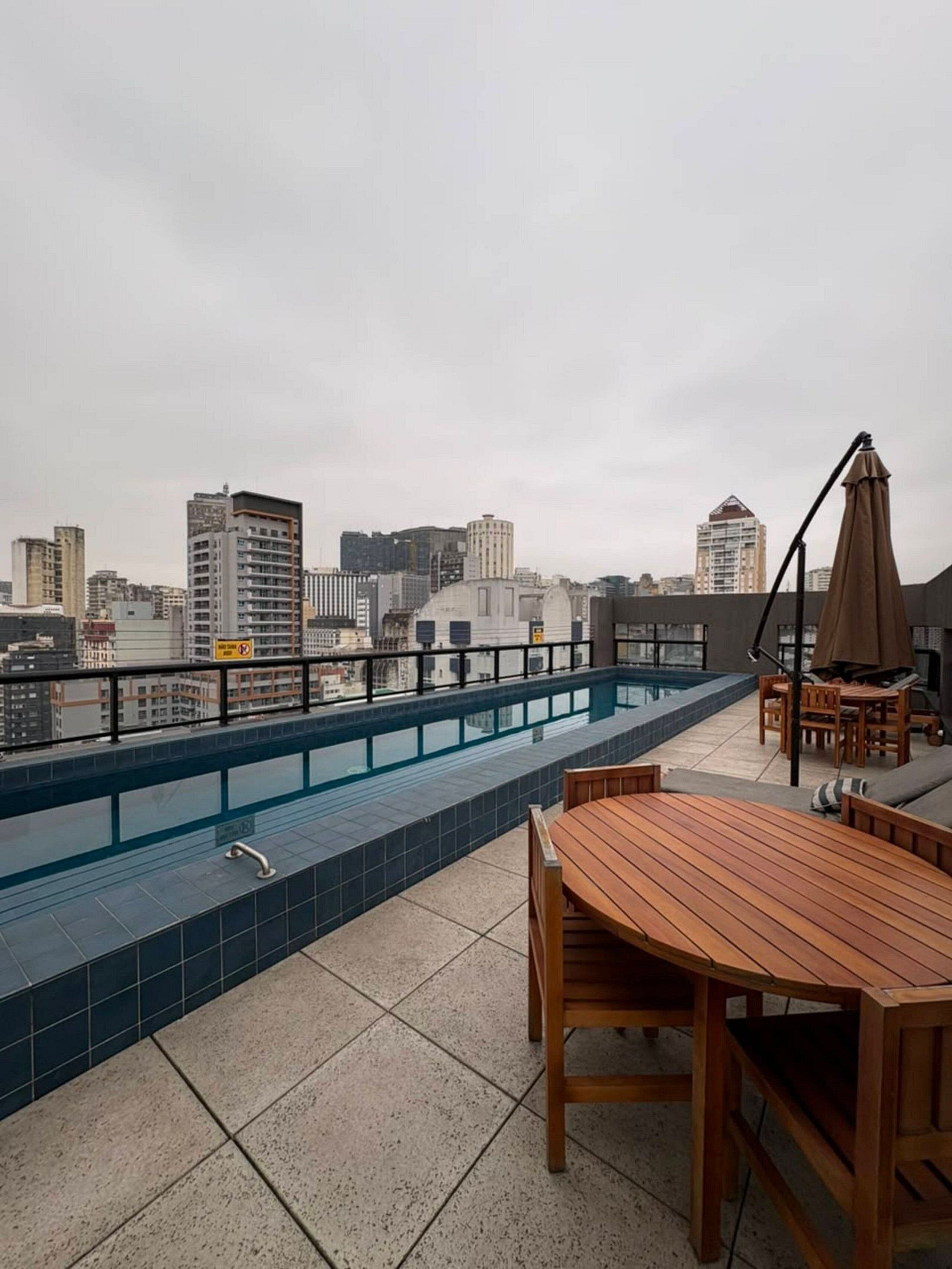 Uma área de piscina no terraço com vista para o horizonte da cidade, uma mesa e cadeiras de madeira e uma espreguiçadeira com guarda-sol.