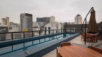 Uma área de piscina no terraço com vista para o horizonte da cidade, uma mesa e cadeiras de madeira e uma espreguiçadeira com guarda-sol.